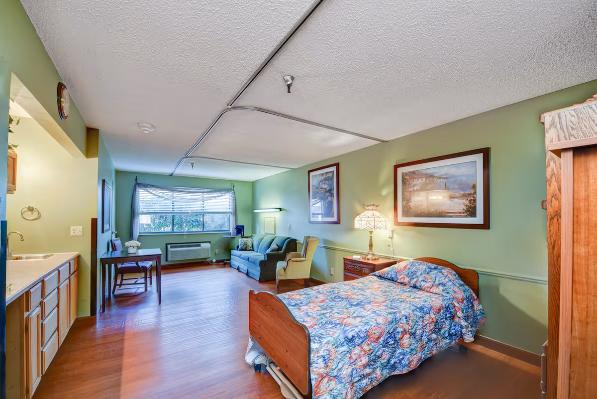 A senior living facility room with a single bed covered in a colorful floral quilt, a wooden nightstand with a decorative lamp, two framed landscape paintings on a green wall, a green armchair, a blue sofa, a wooden table with two chairs near a window with blinds, and a small kitchenette area with a sink and cabinets.