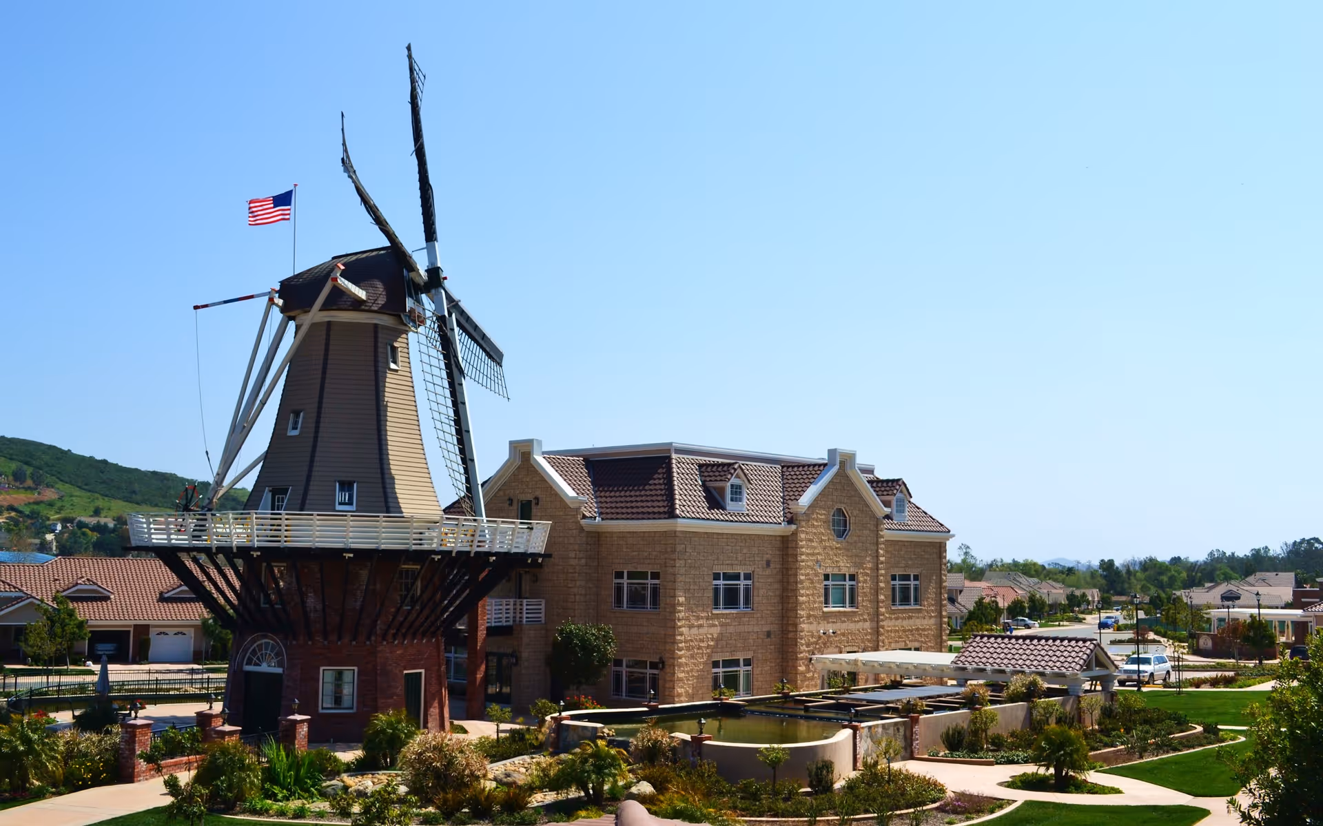 Exterior view of a brick retirement community building with a large decorative windmill, landscaped grounds, and an American flag.