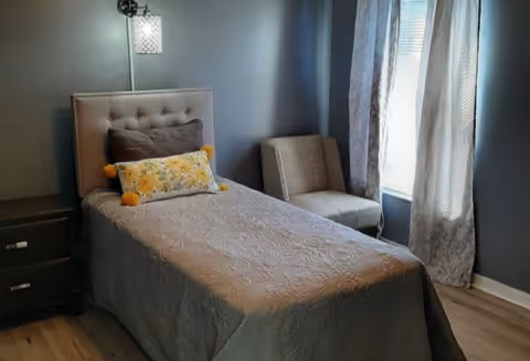 Small bedroom with a single upholstered bed, decorative pillows, nightstand, accent chair, and a window with curtains.