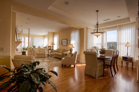 A spacious, well-lit living and dining area with wooden floors, beige walls, and large windows with sheer curtains. The living room features beige sofas, armchairs, a glass coffee table, and decorative lamps. The dining area has a wooden table with upholstered chairs and a hanging light fixture above it. Several plants and framed pictures add to the cozy ambiance.