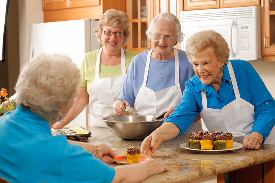 Four elderly women in a kitchen setting, three wearing aprons and smiling as they prepare and serve cupcakes. One woman is handing a cupcake to another seated woman across the counter. The kitchen has wooden cabinets and a microwave in the background.
