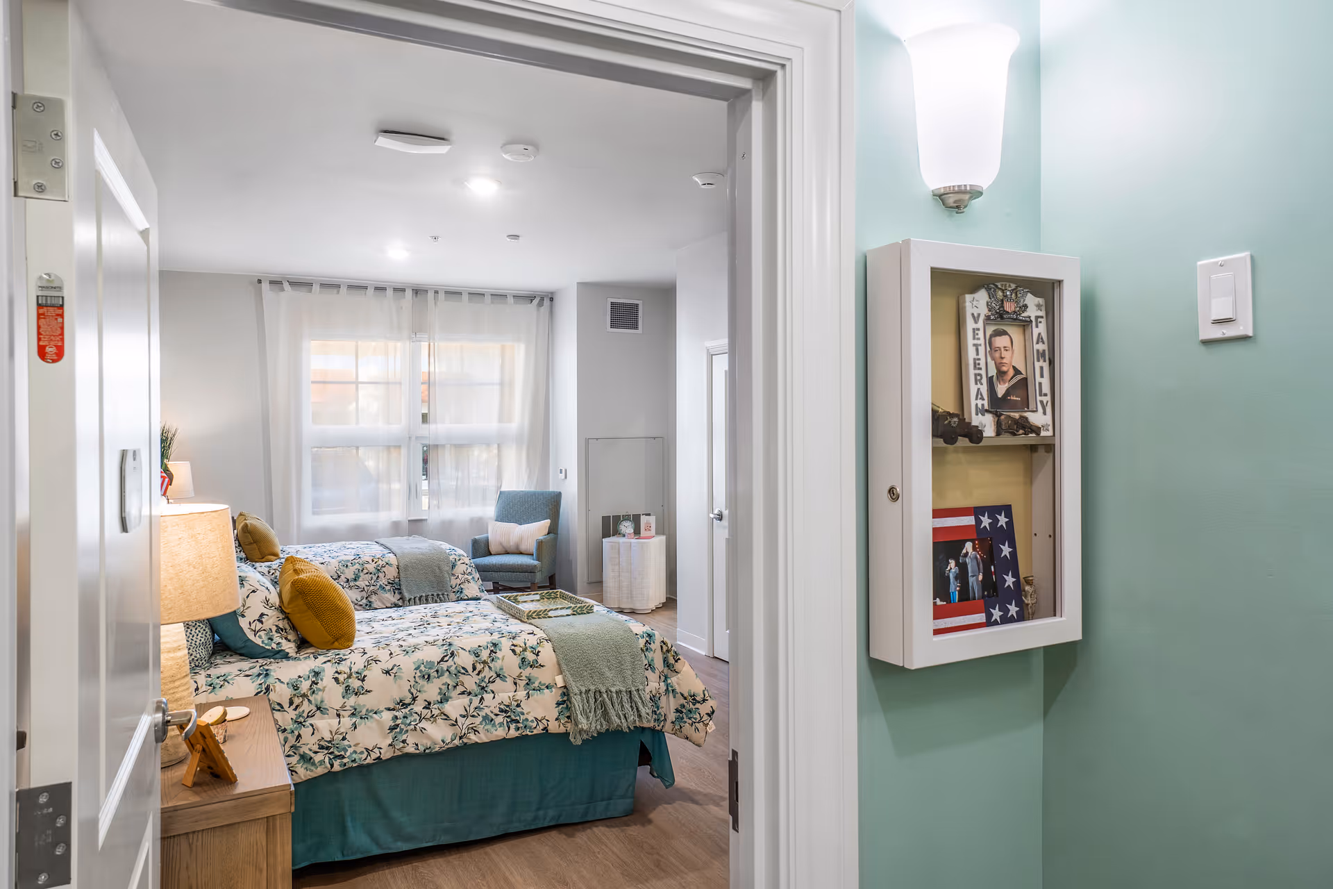 View through a doorway into a furnished memory care bedroom with twin beds, floral bedding, a windowed seating area, and a wall-mounted display case.