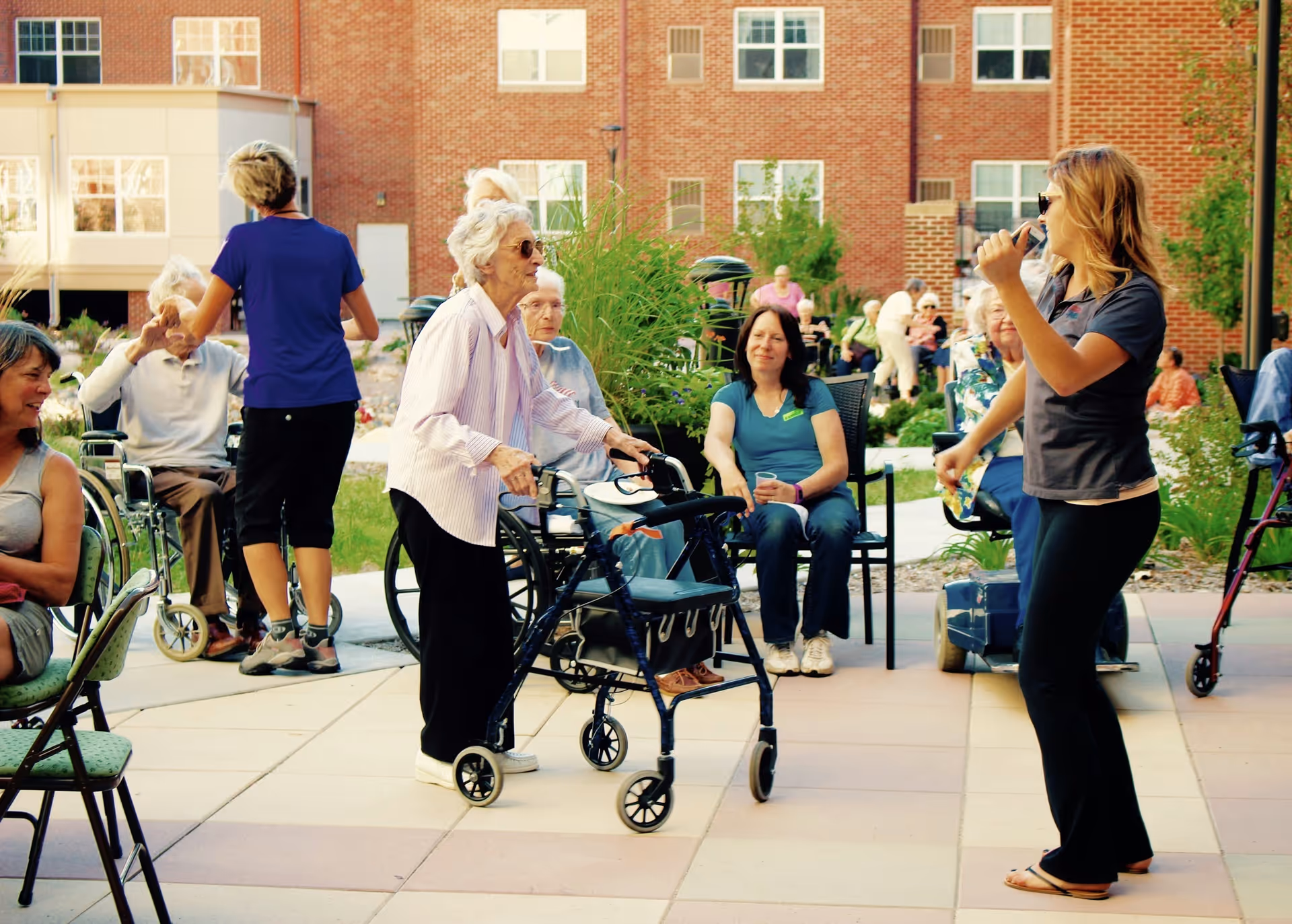 A group of elderly people and caregivers are gathered outdoors on a patio area in front of a brick building. Some seniors are seated, while others use walkers or wheelchairs. A woman in a dark shirt and sunglasses is speaking or singing into a microphone, engaging the group. The scene is lively and social with greenery and paved walkways.