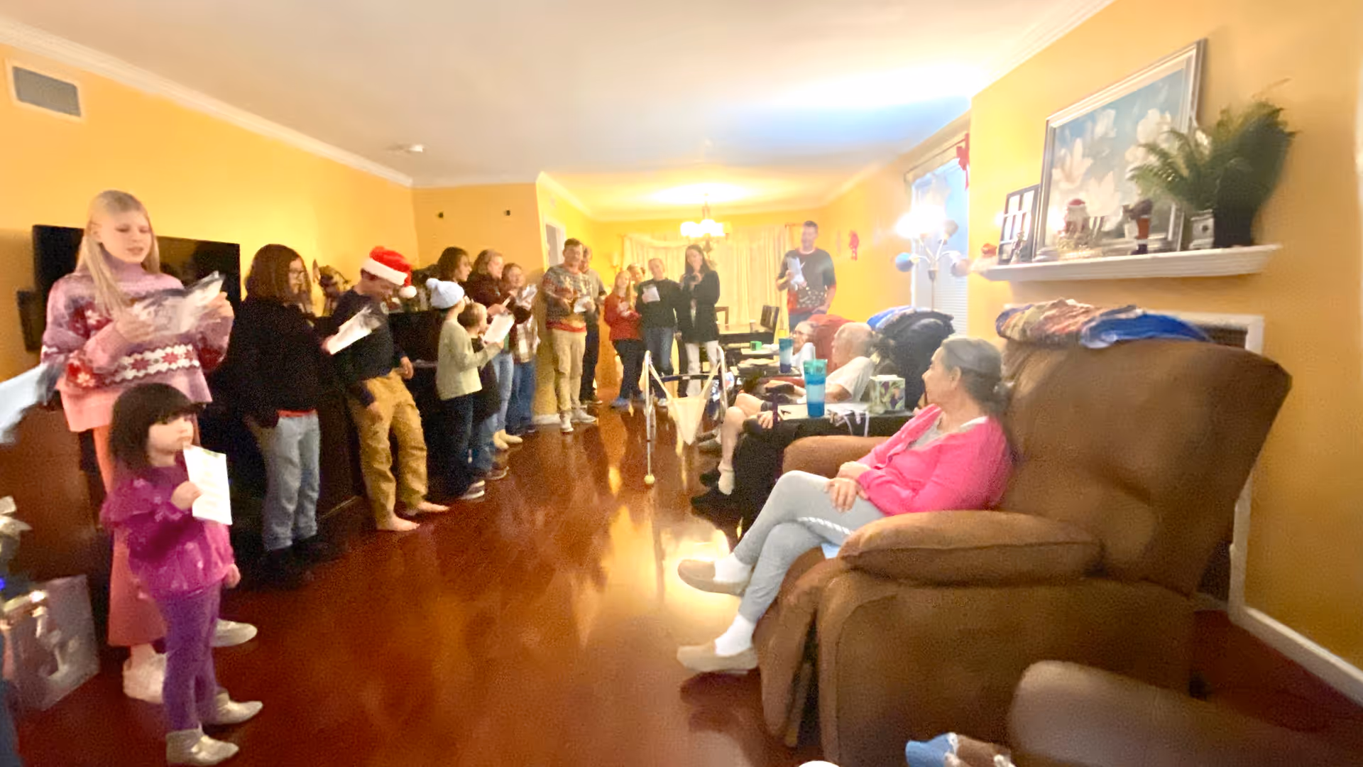 A group of people, including children and adults, standing in a line singing or reading from papers in a warmly lit living room with yellow walls and wooden floors. Several elderly individuals are seated in recliners along one side of the room, watching the group. The room is decorated with framed pictures, plants, and a fireplace mantel.