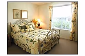 A cozy bedroom with a metal frame bed adorned with floral-patterned bedding and matching pillows. The room has a window with floral curtains allowing natural light to enter, a nightstand with a lamp, and framed artwork on the wall.