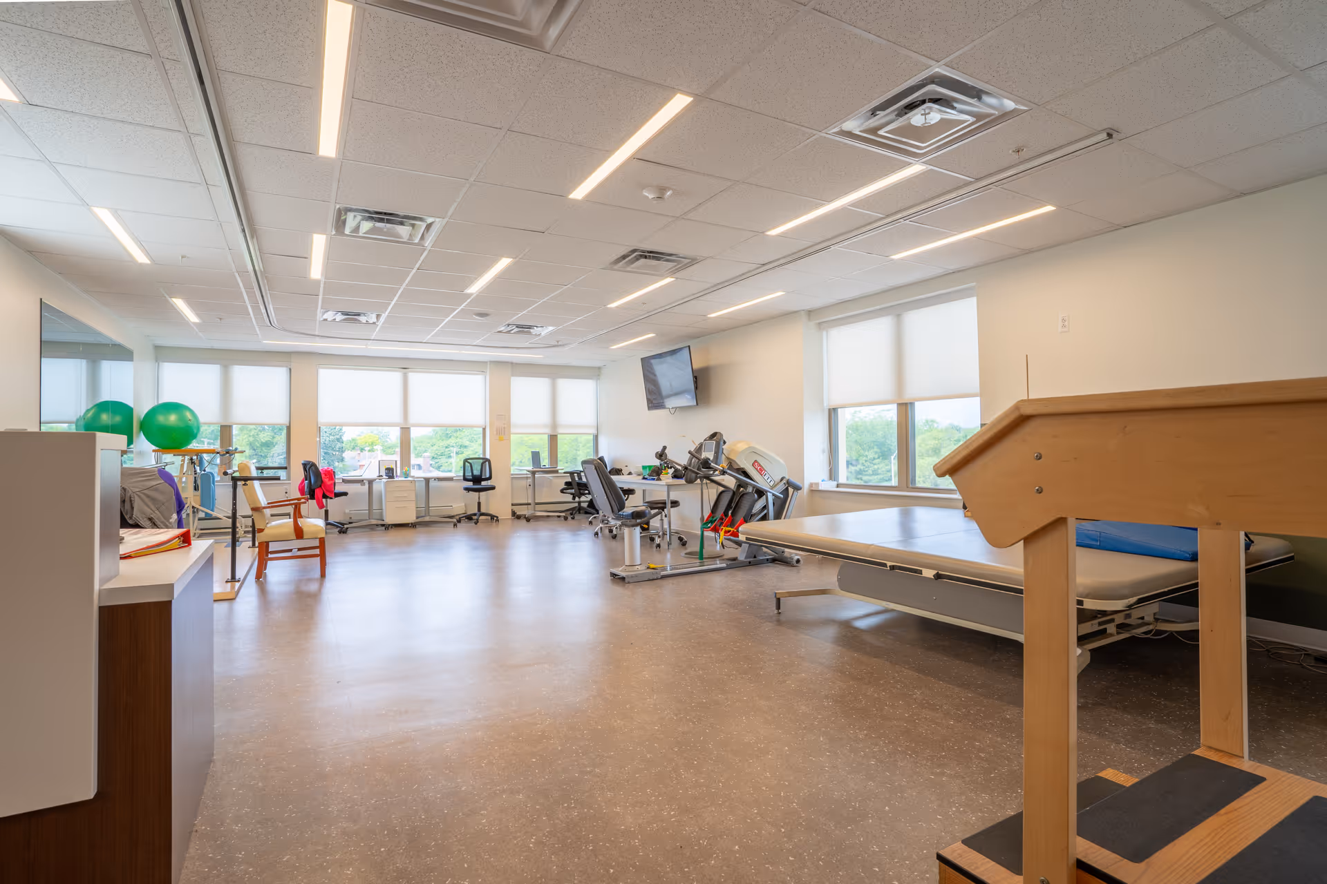 A spacious physical therapy or rehabilitation room with exercise equipment including a treadmill, therapy table, chairs, and green exercise balls near large windows with white blinds. The room has a clean, bright atmosphere with overhead lighting and a wall-mounted TV.
