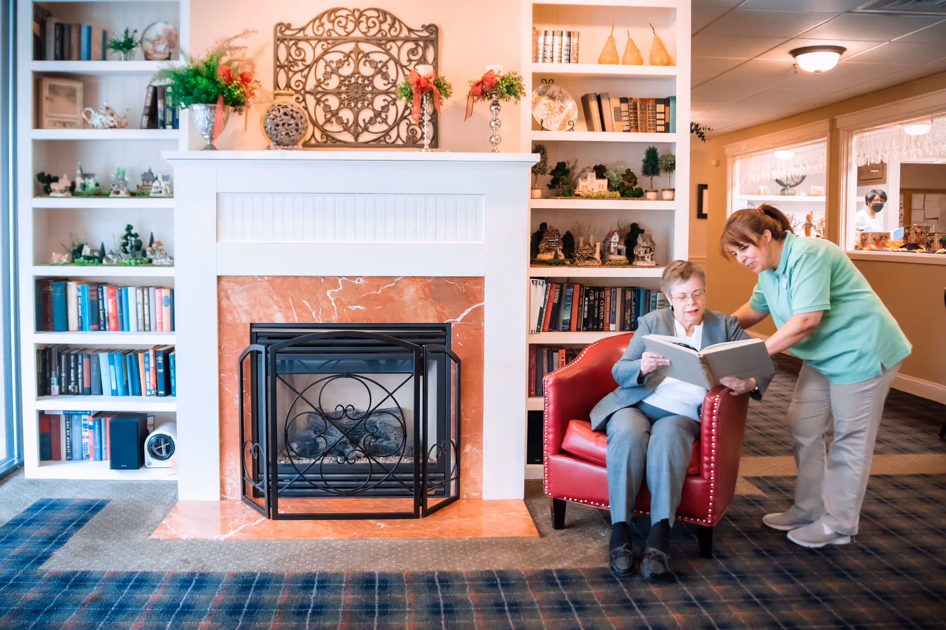 An elderly woman sitting in a red armchair reading a book with the assistance of a caregiver in a green uniform. They are in a cozy room with a fireplace surrounded by built-in bookshelves filled with books and decorative items. The room has a carpeted floor and warm lighting.
