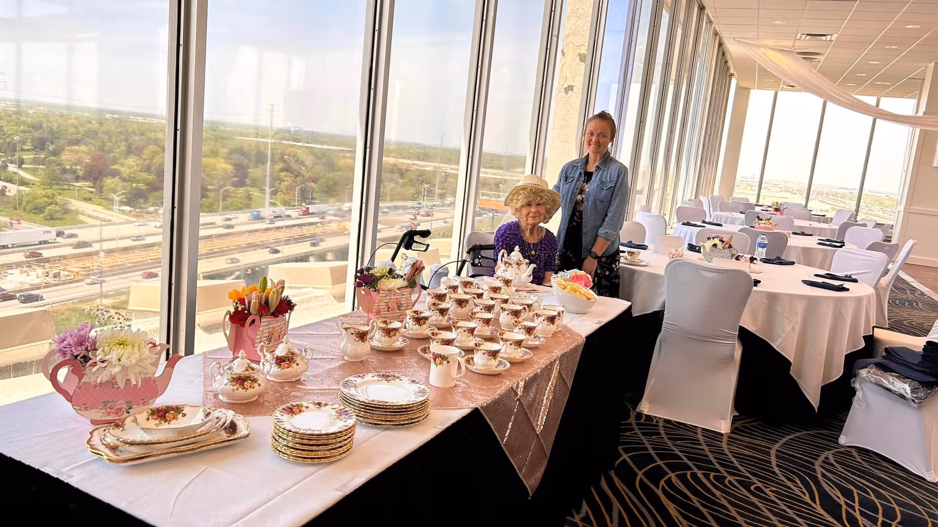 A bright dining room with large windows overlooking a highway and greenery. A table is set with elegant floral china, including teacups, saucers, plates, and teapots. Two women, one elderly seated in a wheelchair wearing a hat and a younger woman standing beside her, are smiling at the camera. Several round tables with white tablecloths and chairs covered in white fabric are arranged in the background.
