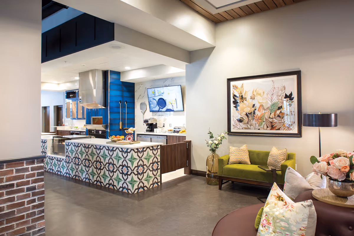 A modern interior space featuring a kitchen area with a patterned tile island and stainless steel appliances, adjacent to a cozy seating area with a green loveseat, decorative pillows, a floor lamp, and a framed floral artwork on the wall.