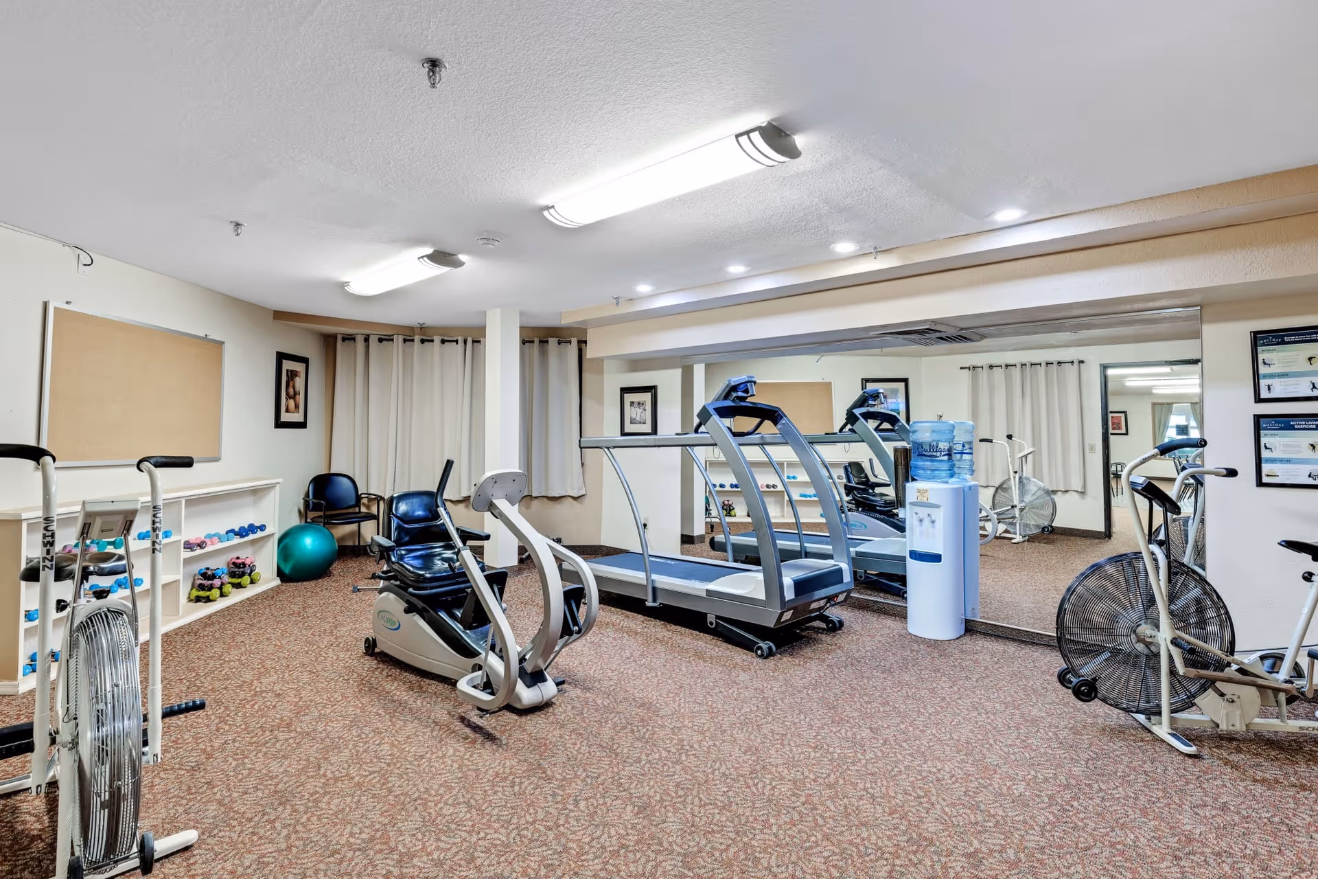 A small fitness room with exercise equipment including a treadmill, stationary bikes, and a recumbent bike. There are shelves with various dumbbells and a green exercise ball. The room has carpeted flooring, beige walls, a large mirror on one wall, and fluorescent ceiling lights. A water cooler is also visible near the mirror.