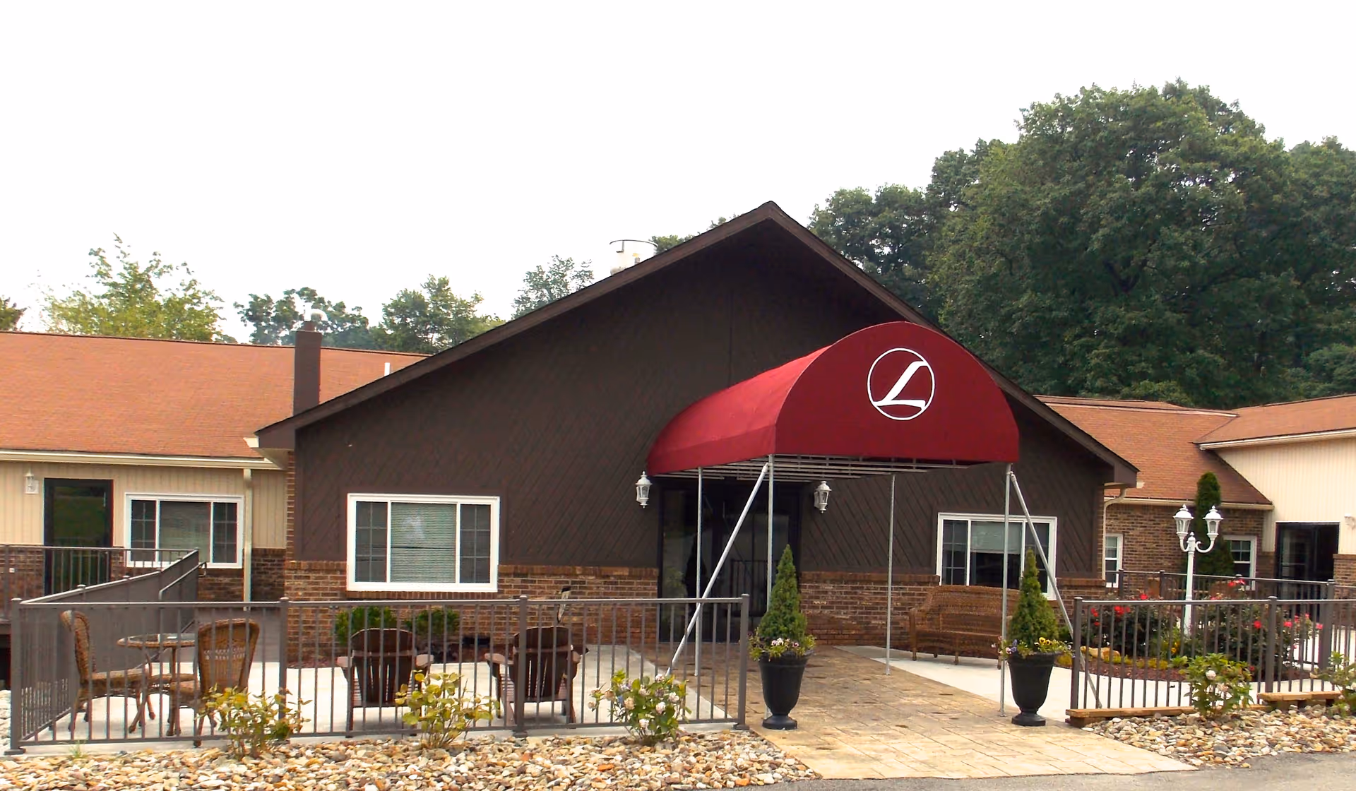 Exterior view of a senior living facility building with a brown and beige facade, a red canopy entrance with a white logo, outdoor seating area with chairs and tables, potted plants, and trees in the background.