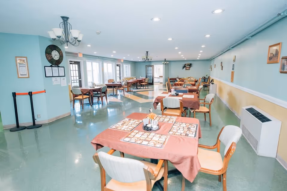 A bright dining room with multiple tables covered in brown tablecloths and set with placemats and condiments. The room has light blue walls, green flooring, and several chairs around each table. There are chandeliers hanging from the ceiling and large windows letting in natural light.
