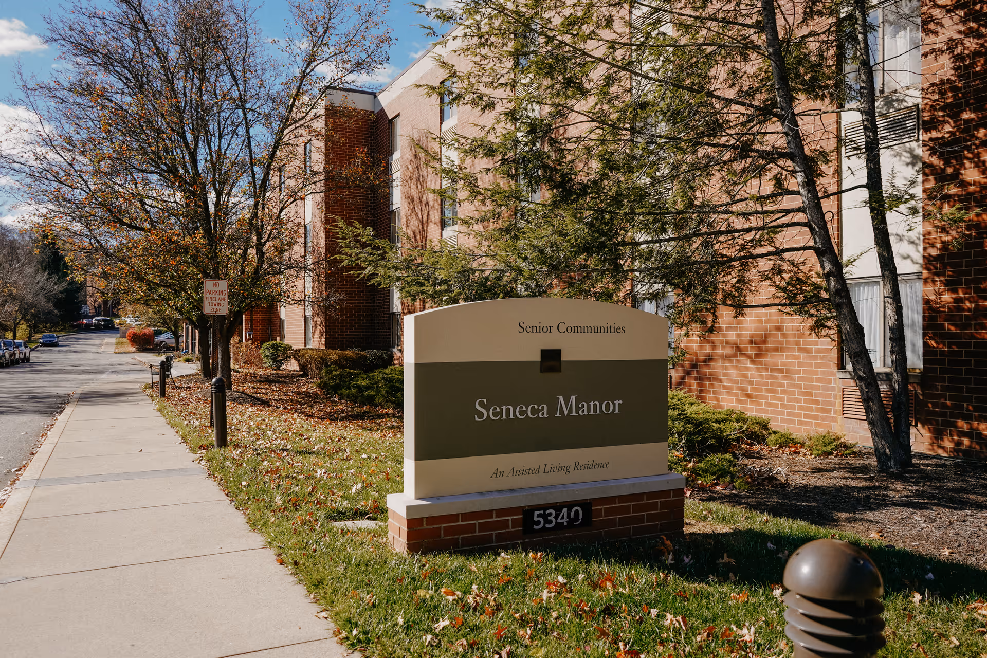 Outdoor view of the entrance area of Seneca Manor, an assisted living residence, showing a sign with the facility name and address 5349. The building is made of red brick and there are trees and a sidewalk along the street.