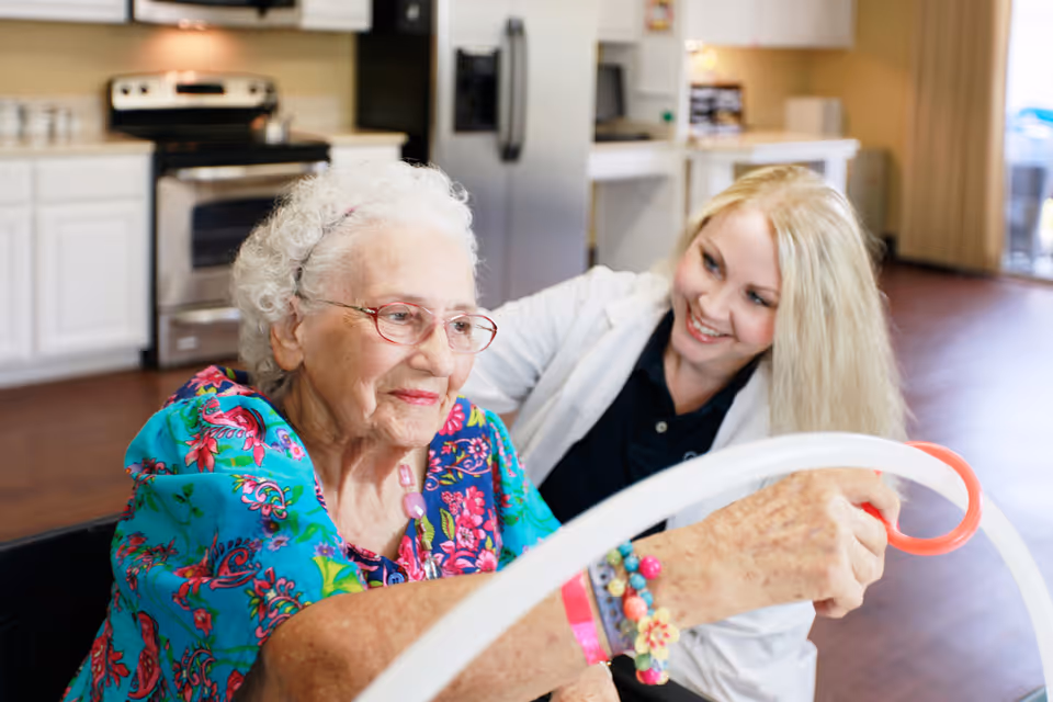 An elderly woman wearing glasses and a colorful floral blouse is engaged in an activity with a smiling caregiver in a white coat in a bright kitchen setting.