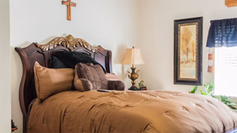 A bedroom with a large wooden bed featuring an ornate headboard. The bed is made with brown bedding and several pillows. There is a bedside table with a decorative lamp and a small figurine. A framed picture hangs on the wall next to a window with a dark blue valance. A wooden cross is mounted on the wall above the bed.