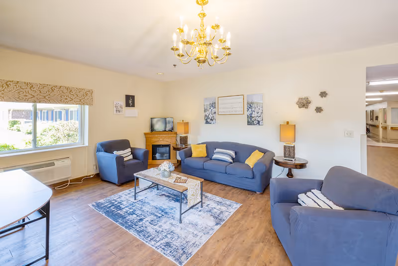 Bright communal living room with blue sofas and armchairs around a coffee table, a TV in the corner, and a chandelier overhead.