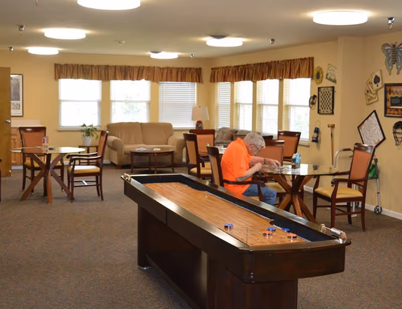 A senior man in an orange shirt sitting at a table playing a game in a well-lit common room with multiple tables and chairs, a shuffleboard table in the foreground, and a beige couch near windows with brown valances.