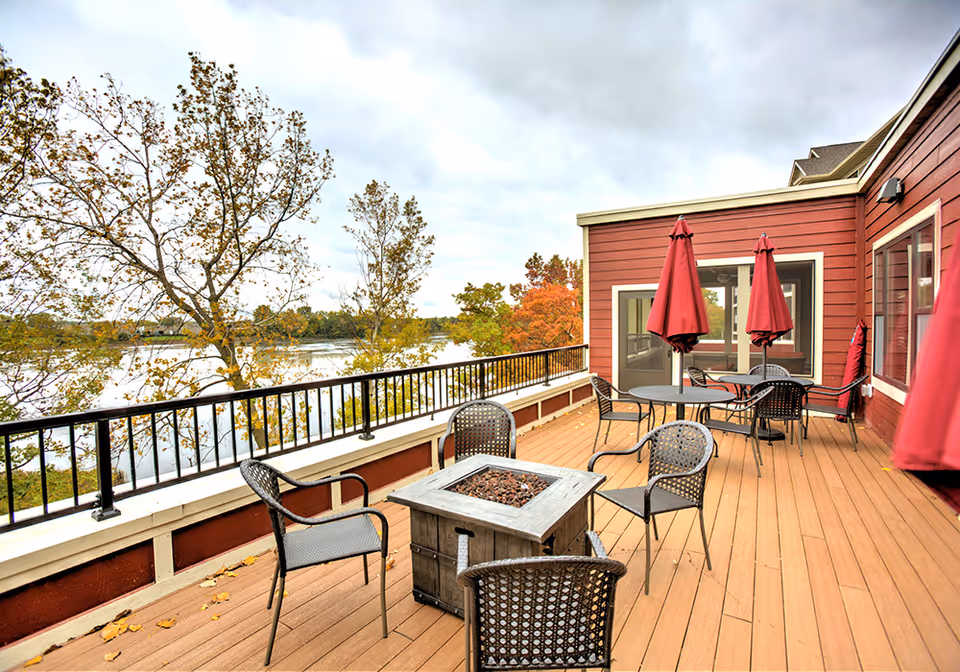 Outdoor patio area at Cardigan Ridge Senior Living with several black metal chairs and tables, some with red umbrellas. The patio overlooks a body of water and trees with autumn foliage under a cloudy sky.