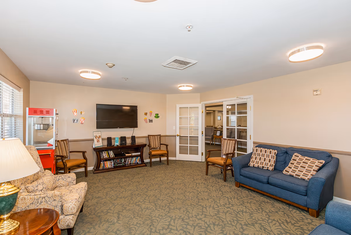A cozy senior living common area with a blue couch, two patterned armchairs, and two wooden chairs. A flat-screen TV is mounted on the wall above a wooden media console filled with books and DVDs. A red popcorn machine is positioned near the window, and the room is lit by ceiling lights and a table lamp.