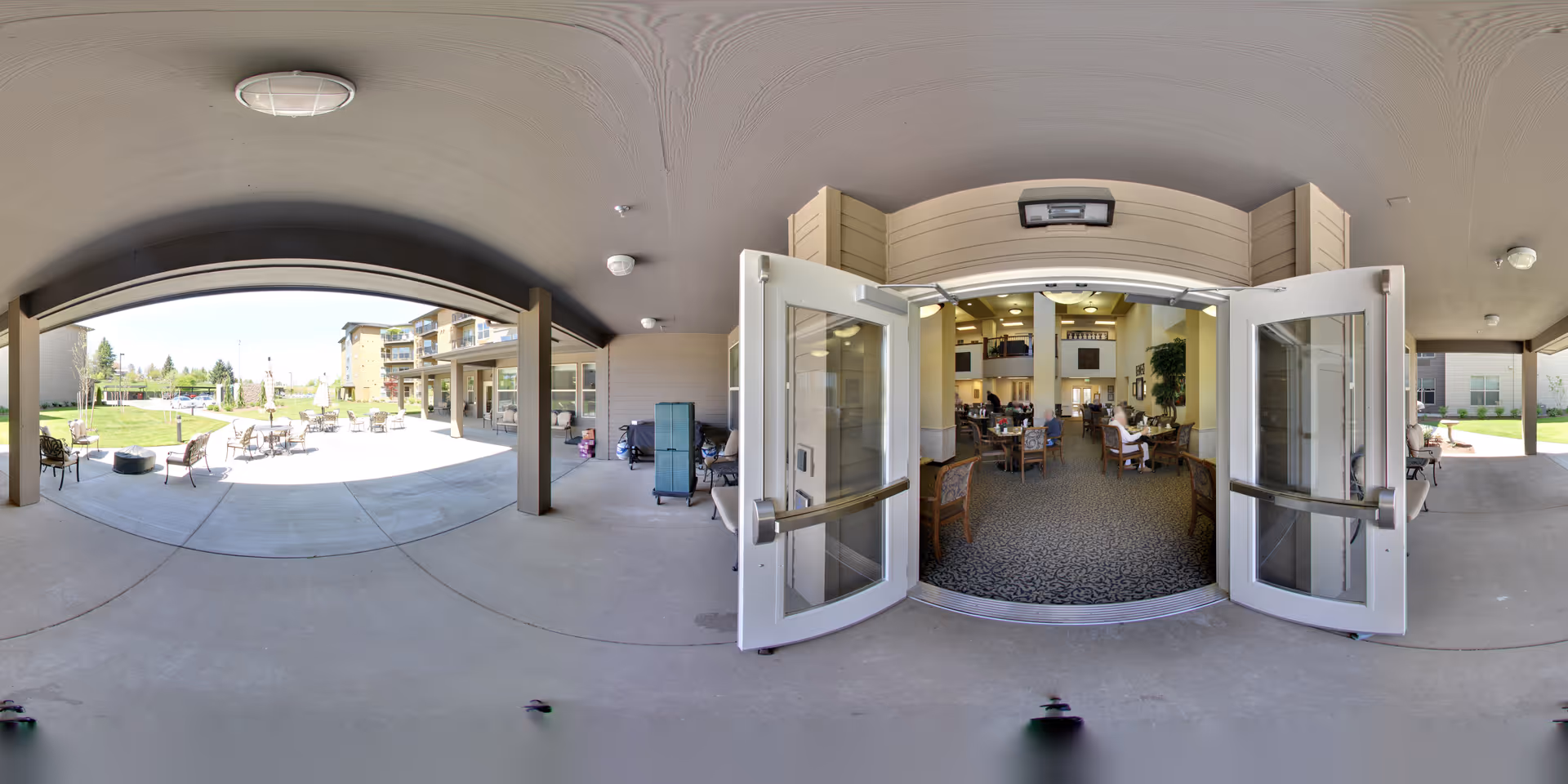 View from a covered patio area looking through open double doors into a spacious indoor common area with tables and chairs where people are seated. Outside, there are more tables and chairs on a concrete patio with a grassy lawn and buildings in the background under a clear sky.