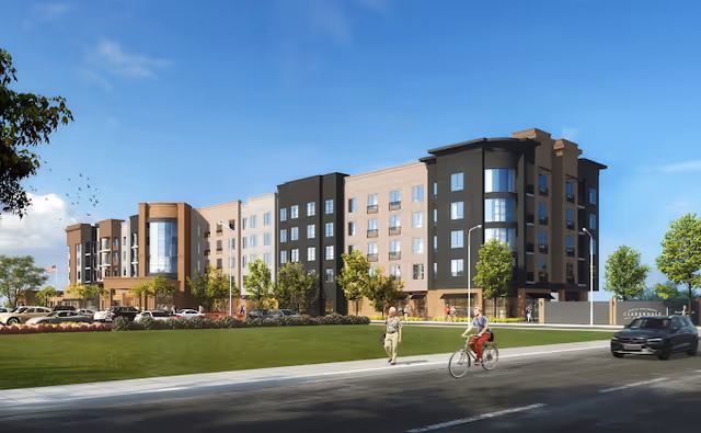 Modern multi-story senior living facility exterior with landscaped lawn, cars, pedestrians, and cyclists.