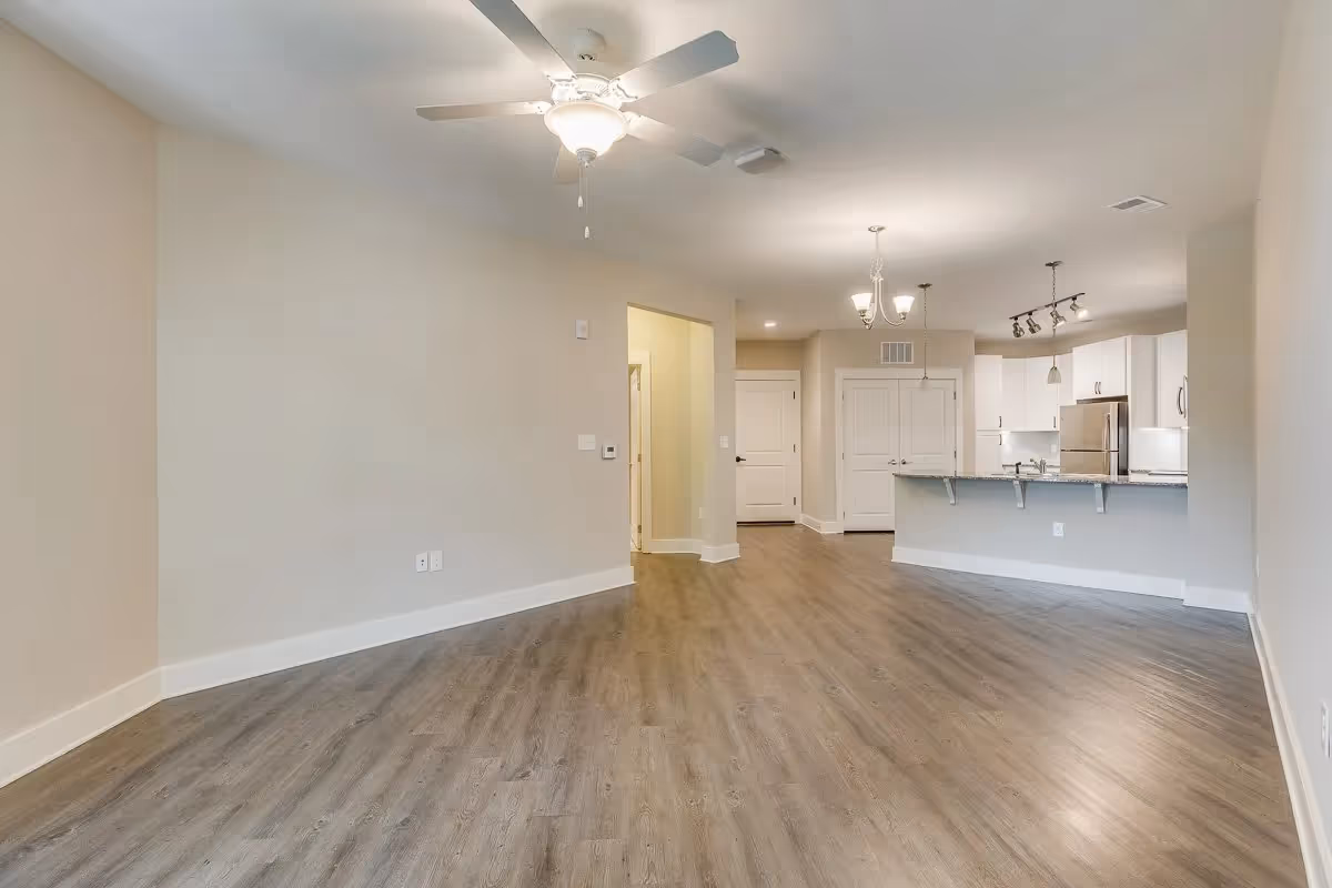Open living area with a ceiling fan, wood-look flooring, and a kitchen with a breakfast bar and stainless appliances in the background.