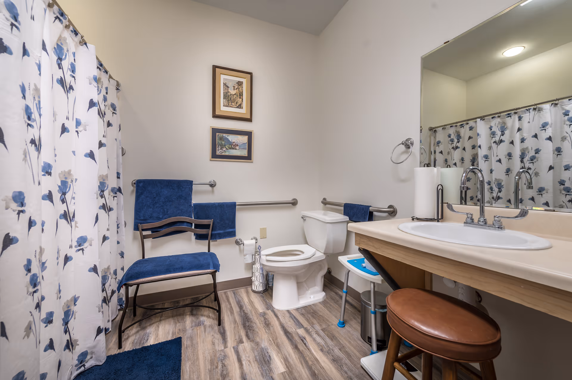 A clean and accessible bathroom with a white toilet, a sink with a large mirror above it, a wooden stool, and a chair with blue cushions. The shower has a white curtain with blue floral patterns. There are two framed pictures on the wall and blue towels hanging on metal grab bars. The floor has wood-like vinyl flooring.