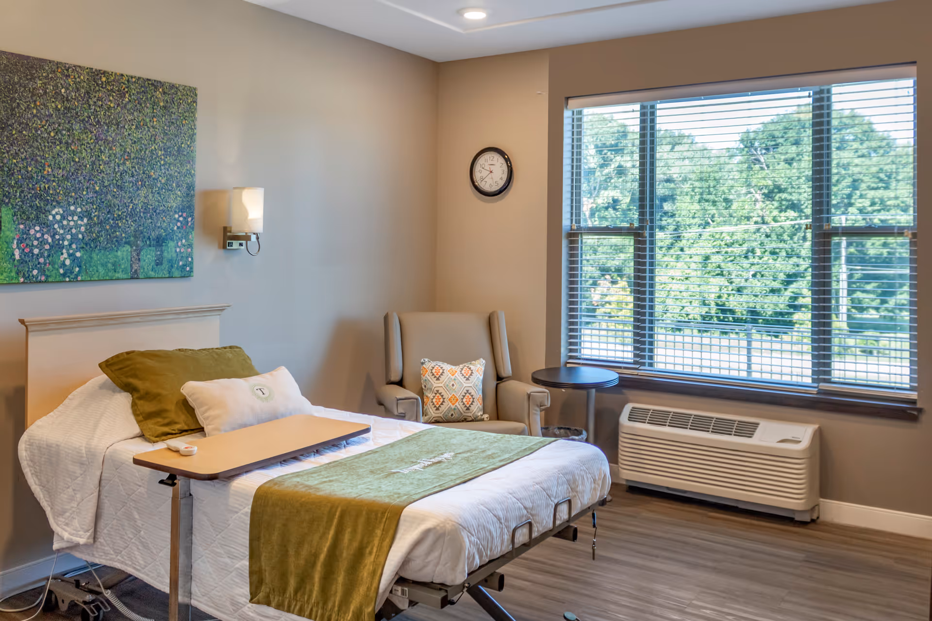 A well-lit room with a hospital-style bed covered with white and green bedding, a green pillow, and a beige pillow. There is a small overbed table on the bed. Next to the bed is a beige armchair with a decorative pillow and a small round side table. A large window with blinds shows green trees outside. The walls are painted beige, with a colorful painting and a wall clock visible.