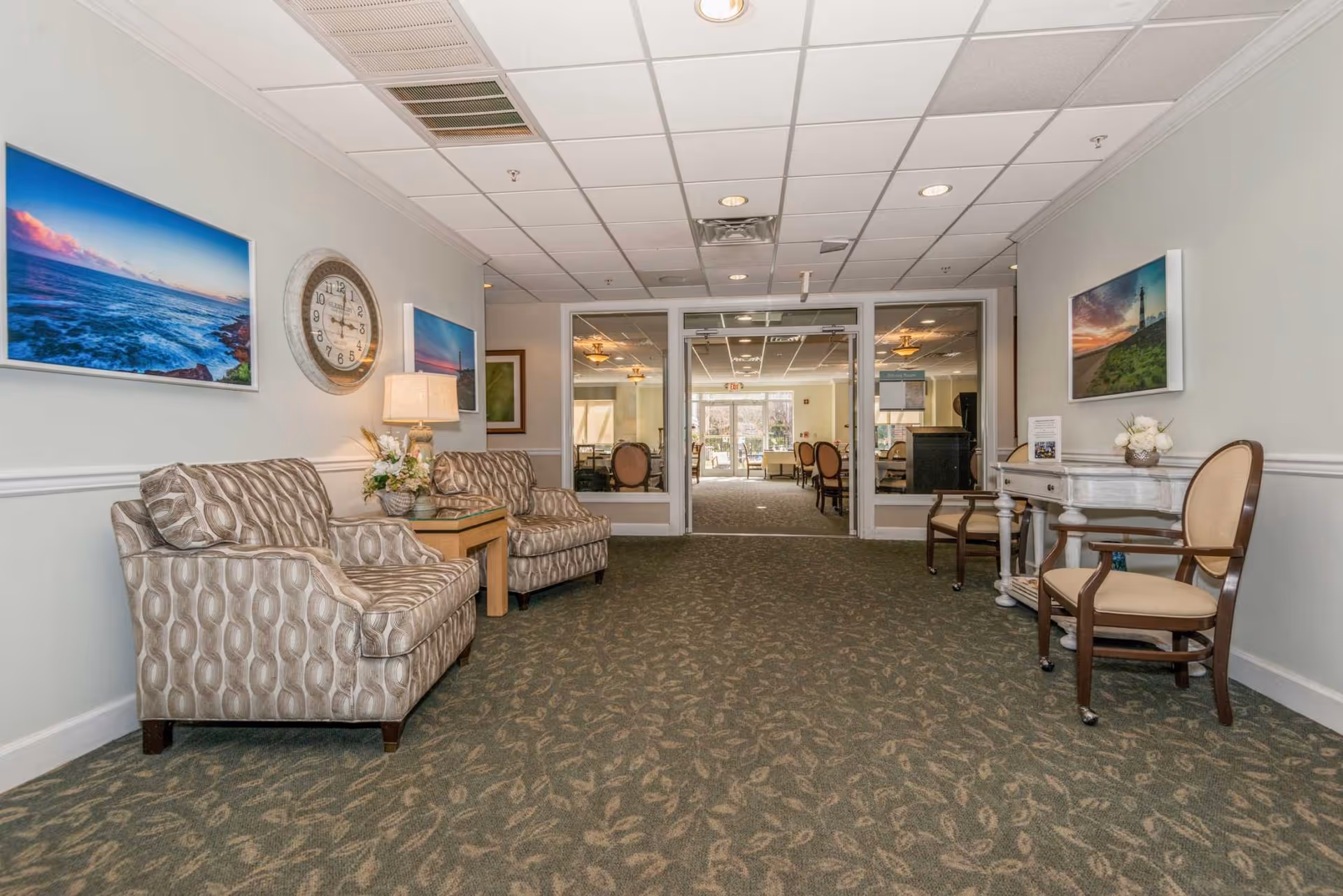 A hallway in a senior living facility with patterned armchairs on the left and wooden chairs with beige cushions on the right. There is a small table with a lamp and flowers between the armchairs, and a white console table with flowers on the right. The walls are decorated with framed pictures of ocean and lighthouse scenes, and a large round clock. At the end of the hallway, glass double doors lead to a dining room with tables and chairs visible.