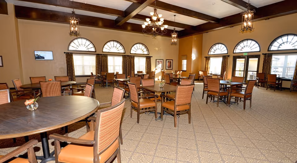 A spacious dining room with multiple round and square tables surrounded by wooden chairs with cushioned seats. The room features large arched windows with curtains, beige walls, and a patterned carpet. Elegant chandeliers and lantern-style light fixtures hang from the ceiling with wooden beams.