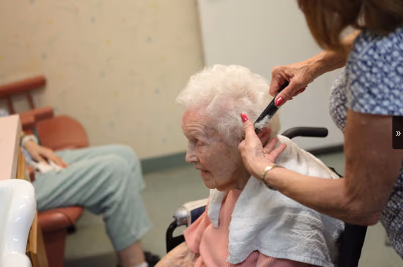 An elderly woman with white hair is seated in a wheelchair while another person combs her hair. The elderly woman has a towel draped around her shoulders. In the background, another person is seated on a chair with legs crossed.