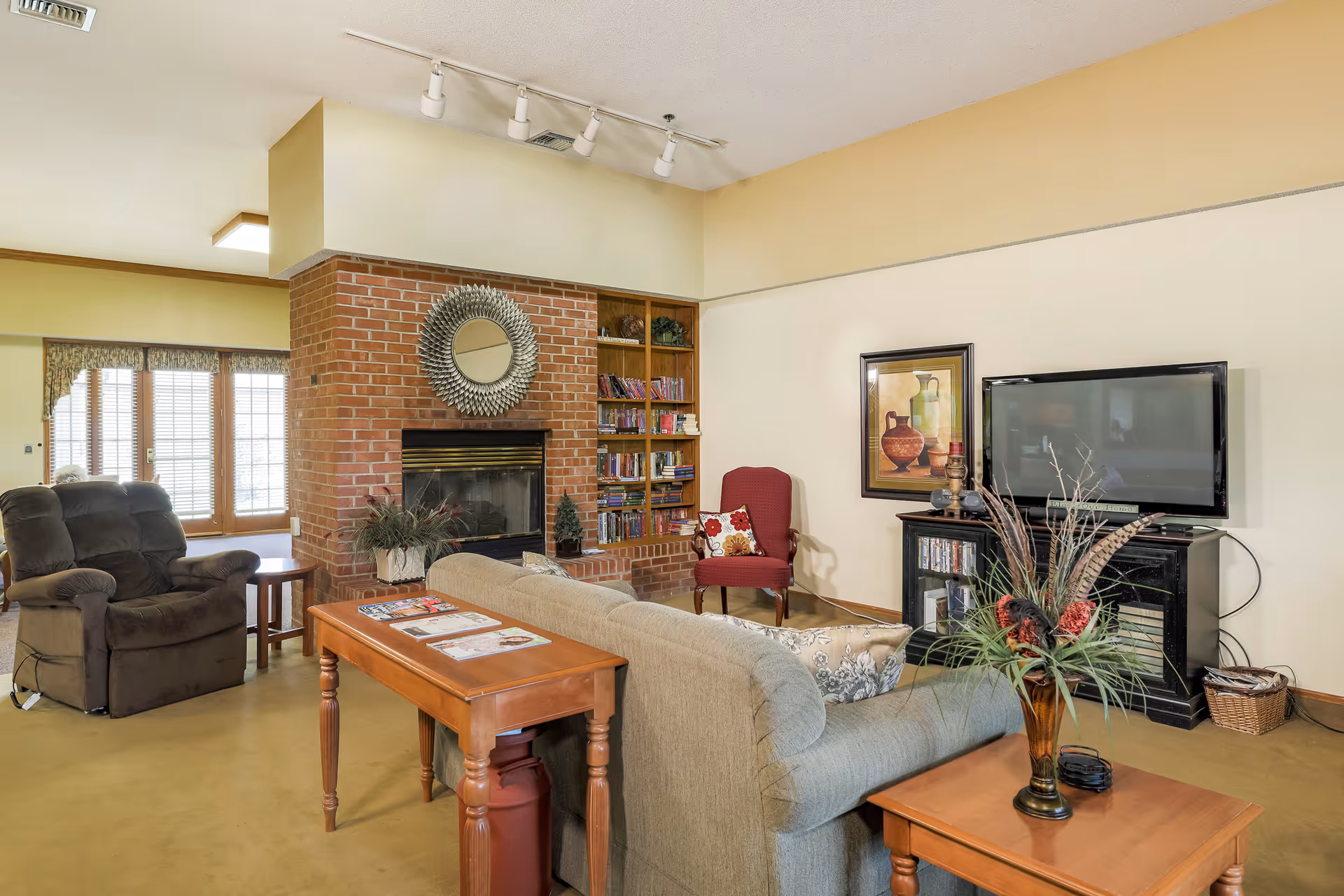 A cozy living room in a senior living facility featuring a brick fireplace with a decorative round mirror above it, a built-in bookshelf filled with books, a beige couch with patterned pillows, a red armchair, a dark recliner chair, a wooden coffee table with magazines, a TV on a black stand, and a vase with decorative plants on a side table.