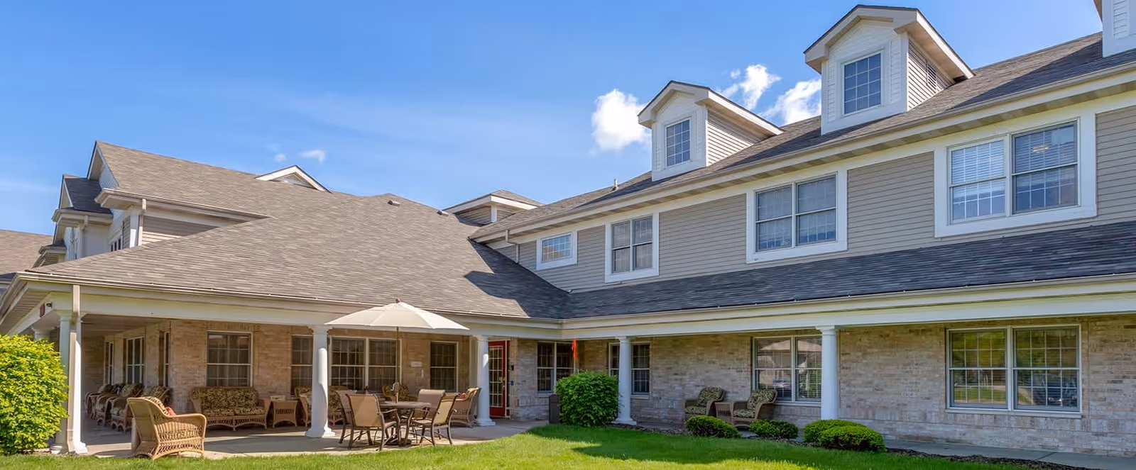 Exterior view of a senior living facility building with beige siding and brick walls, featuring a covered patio area with outdoor seating including chairs, a table, and an umbrella, surrounded by green grass and bushes under a blue sky with a few clouds.