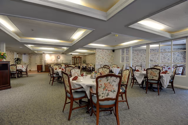 A spacious dining room with multiple round tables covered with white tablecloths and set with napkins and tableware. The chairs have floral upholstery and wooden frames. The room features floral wallpaper, carpeted floors, and recessed ceiling lighting.
