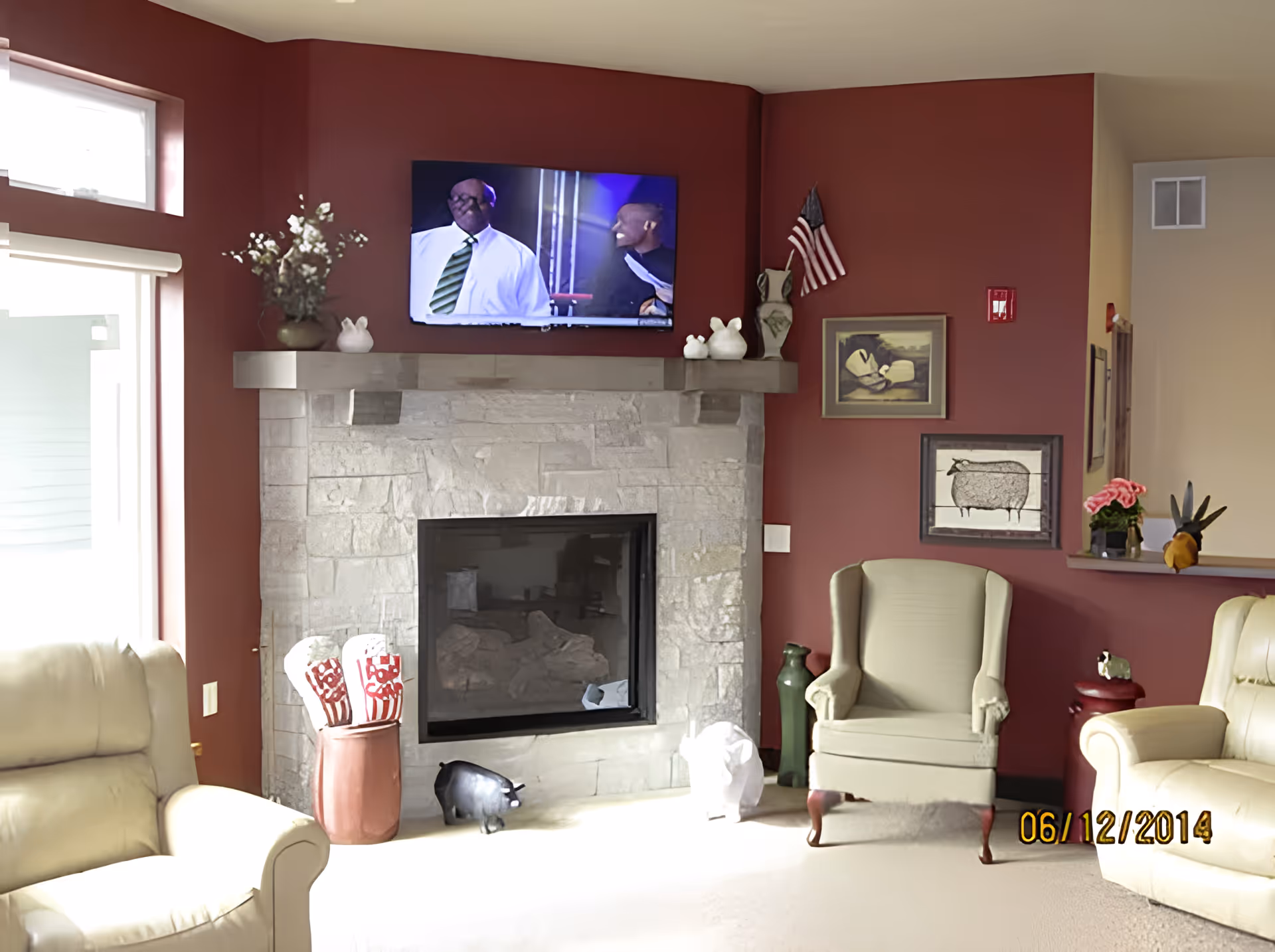 A cozy living room with red walls featuring a stone fireplace with a mounted flat-screen TV above it. The room has beige armchairs, decorative vases, small animal figurines, and framed artwork on the walls. There is a window with a white blind on the left side letting in natural light.