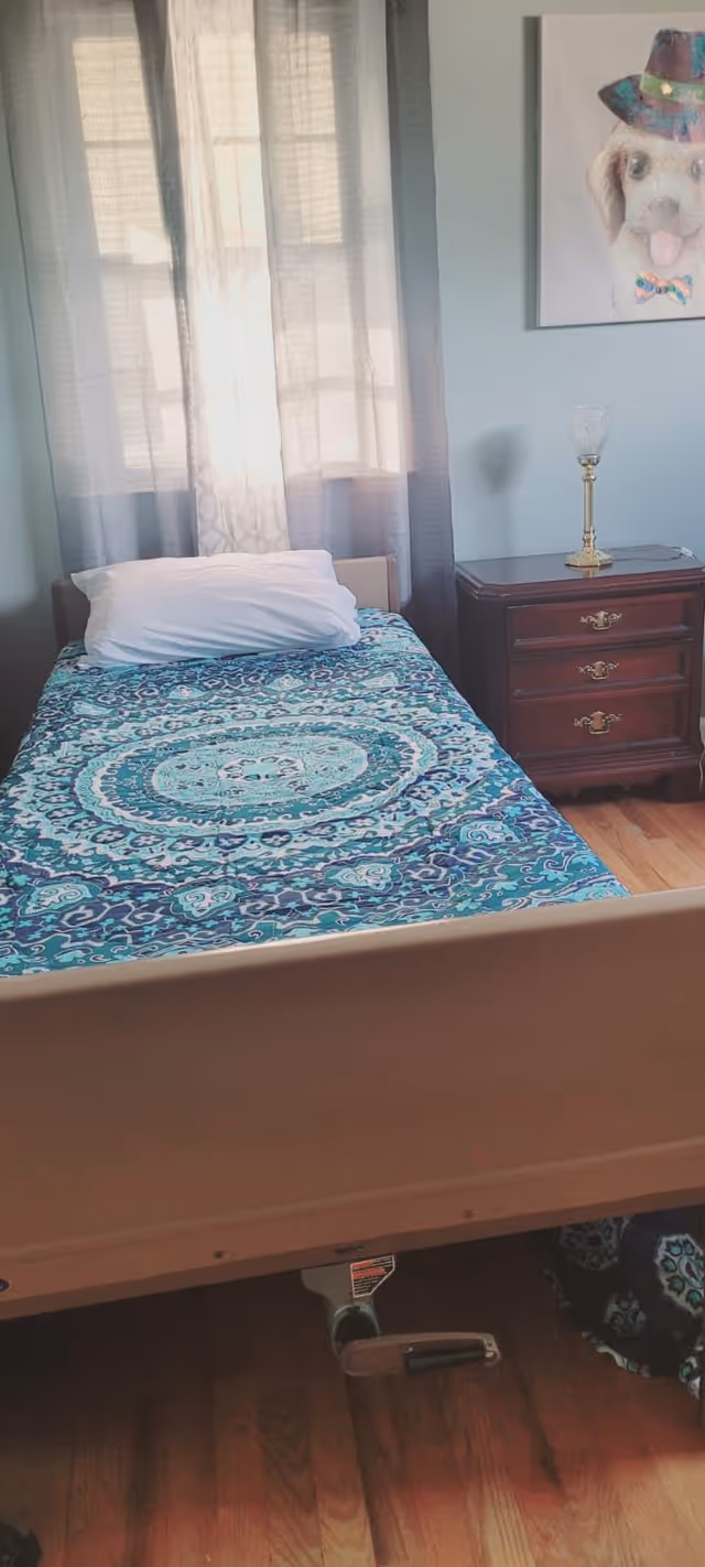 A single bed with a blue and green patterned bedspread and a white pillow is positioned in front of a window with sheer curtains. Next to the bed is a wooden nightstand with three drawers and a decorative glass candle holder on top. A framed picture of a dog wearing a hat and bow tie hangs on the light blue wall above the nightstand. The floor is wooden.