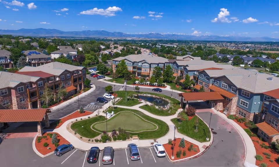 Aerial view of Keystone Place At Legacy Ridge senior living facility showing multiple residential buildings surrounding a landscaped circular driveway with a small pond and putting green in the center. Cars are parked in the parking lot, and mountains are visible in the background under a blue sky with scattered clouds.