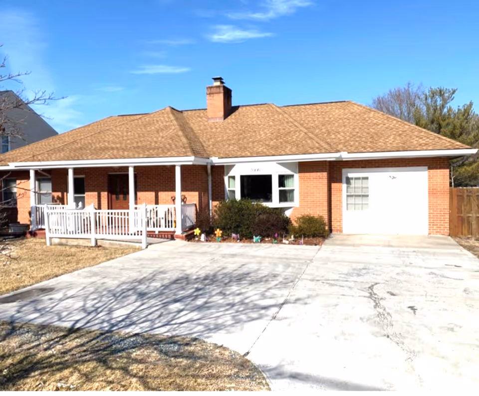 Front exterior view of a single-story brick house with a brown shingle roof, a chimney, a white porch railing, and a concrete driveway. There are bushes and small decorative items in front of the house, and a clear blue sky above.