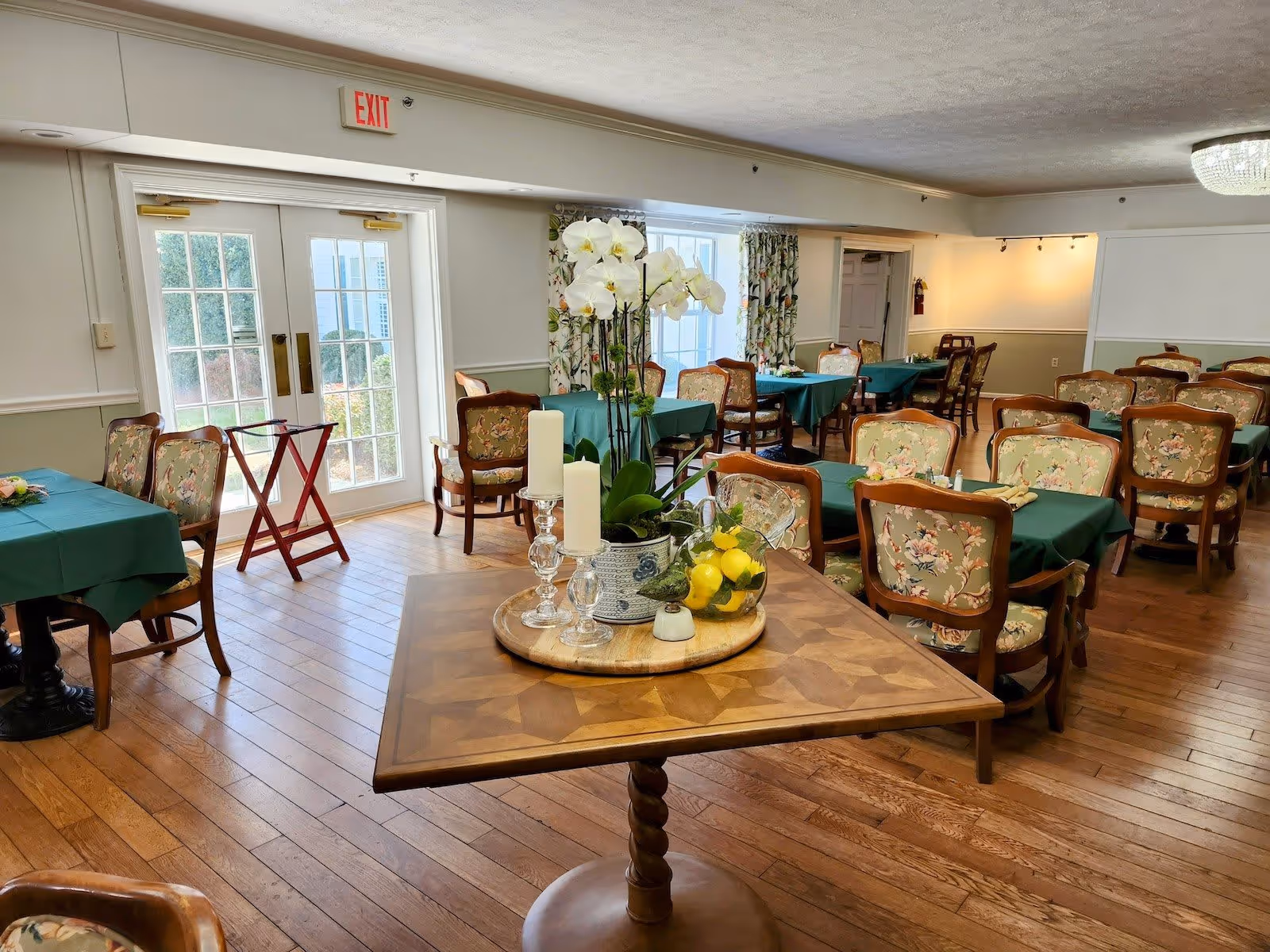 Bright dining room with multiple tables covered in green tablecloths, floral upholstered chairs, and a centerpiece table holding orchids and candles.
