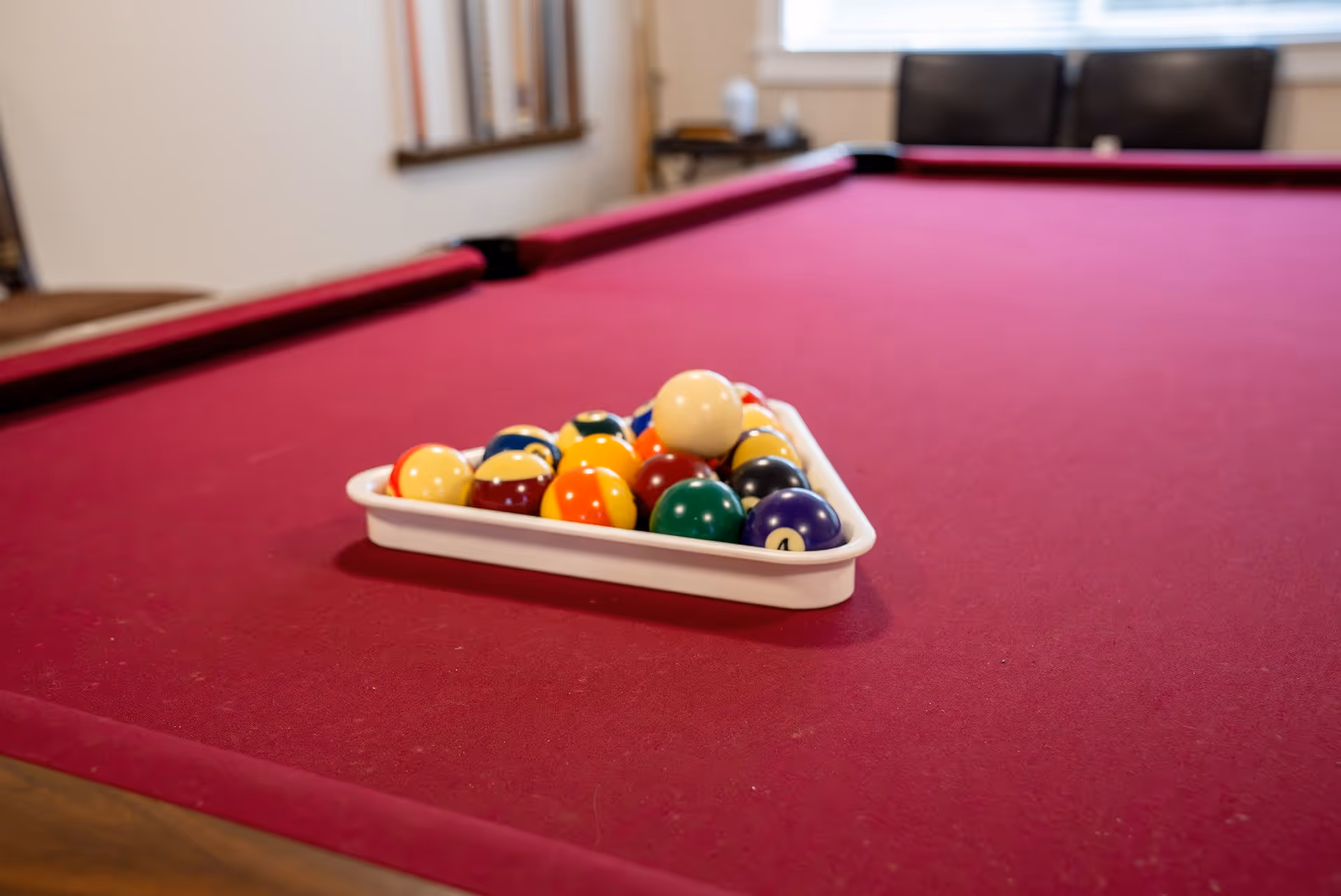 A close-up view of a red felt pool table with a white triangular rack holding a full set of colorful billiard balls, positioned in the center of the table. In the background, a window with blinds and part of a chair are visible.