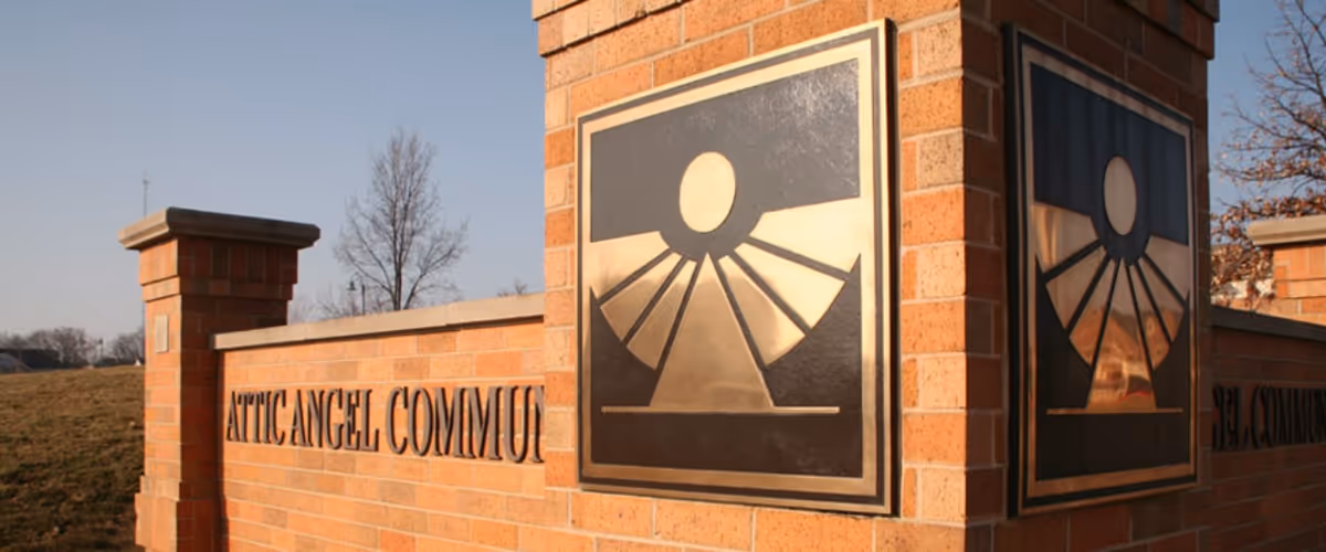 Brick entrance sign for Attic Angel Community with a metal emblem featuring a stylized sun and rays mounted on a brick pillar, set against a clear sky and some bare trees in the background.