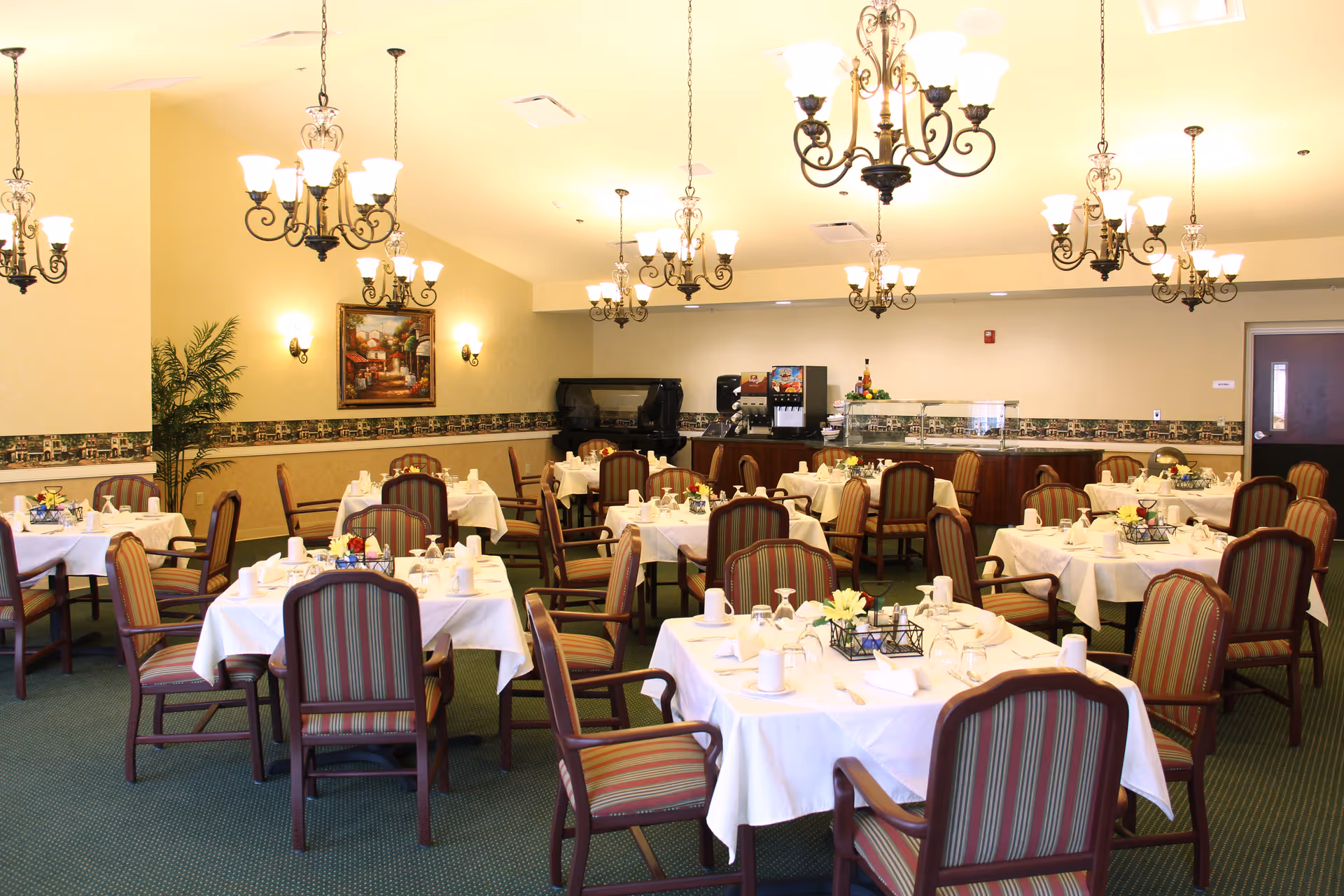 Well-lit dining room with multiple round tables set with white tablecloths and striped chairs under chandeliers.