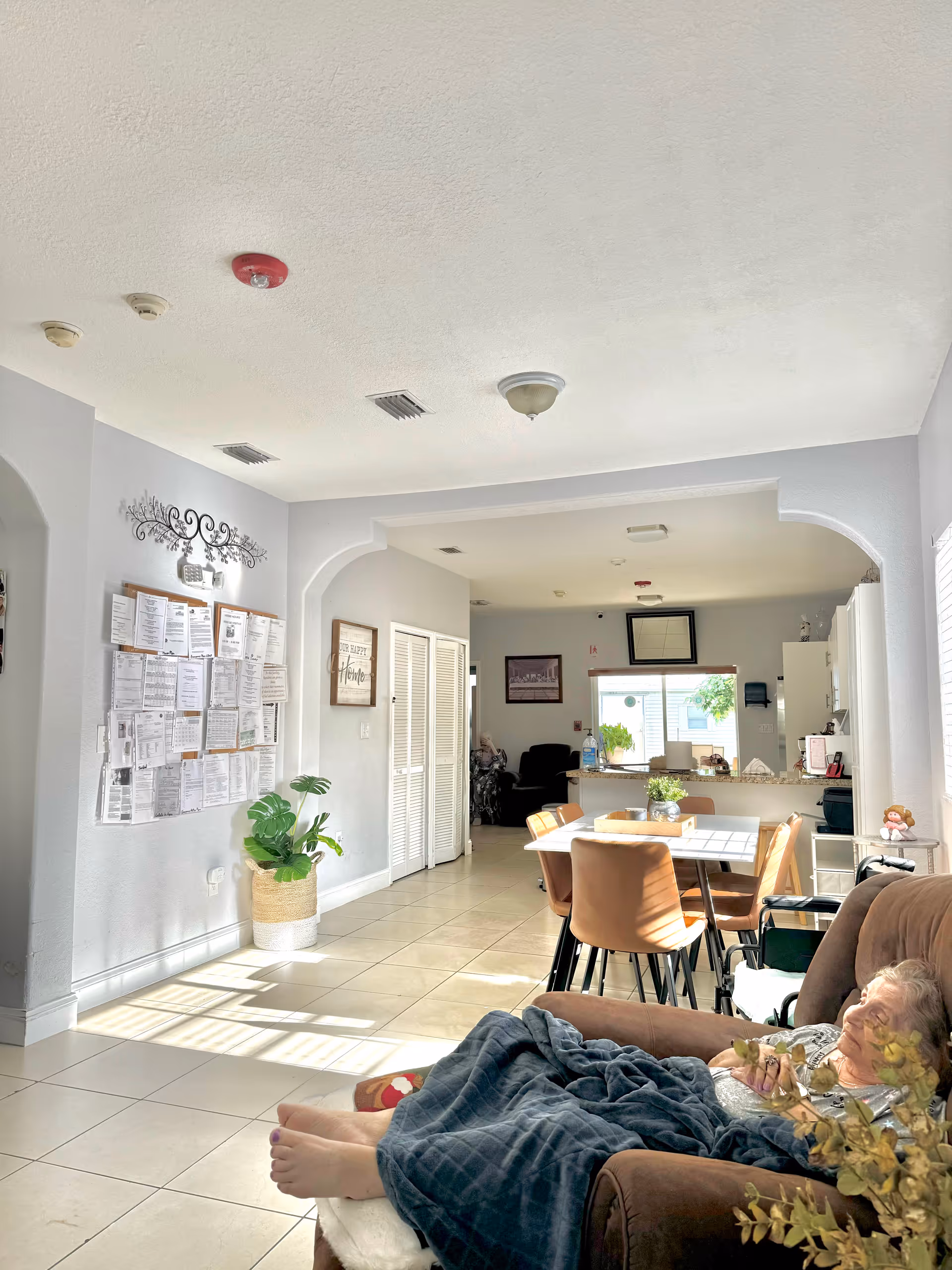 Open-plan living area with a reclining resident in a chair in the foreground, a dining table and chairs in the center, and a kitchen and bulletin board in the background.