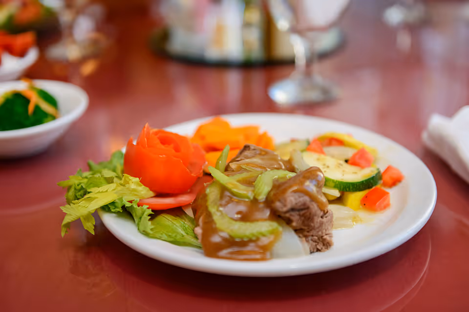 A white plate with a serving of meat topped with brown gravy and celery, accompanied by cooked mixed vegetables including zucchini, carrots, and red bell peppers, and a garnish of lettuce and a tomato rose on a red table.