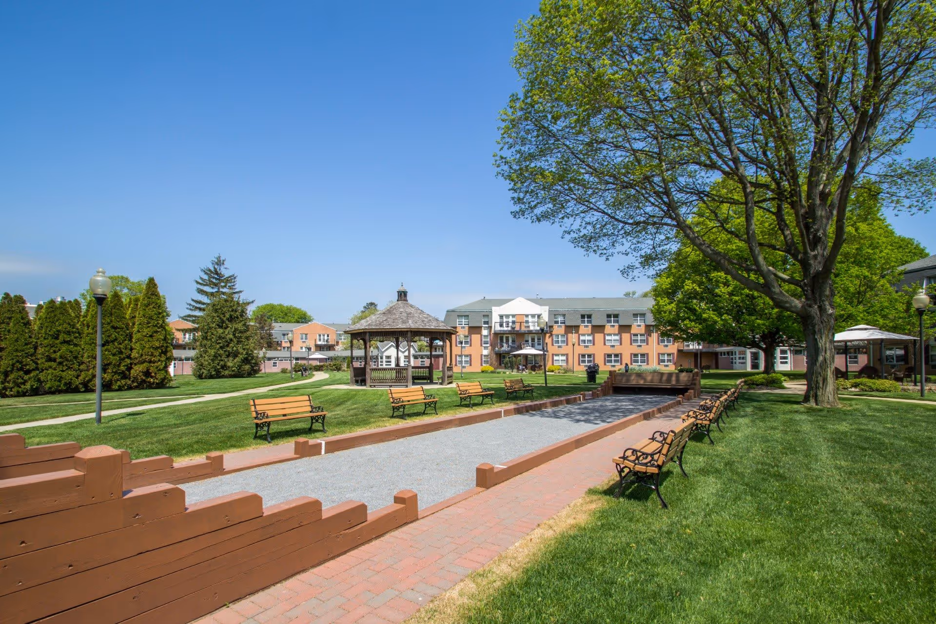 Outdoor view of Dominican Village showing a bocce ball court with benches on both sides, a gazebo in the background, well-maintained green lawns, trees, and a multi-story residential building under a clear blue sky.