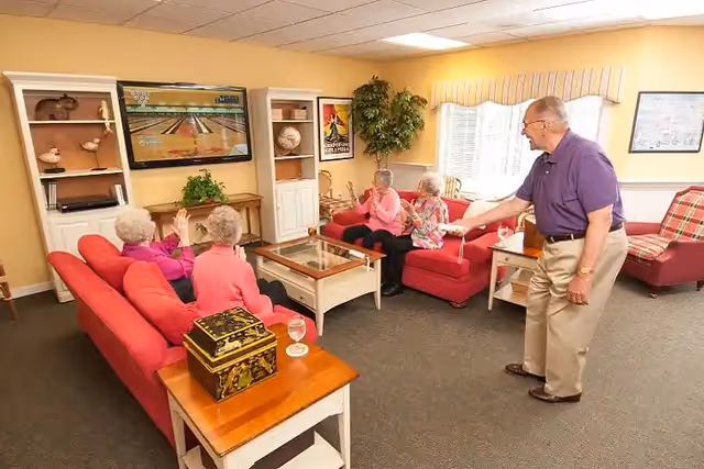 A group of elderly people in a cozy living room setting playing a video bowling game on a large TV mounted on the wall. Four women are seated on red couches clapping and watching, while an elderly man stands holding a game controller, appearing to take a turn. The room has warm yellow walls, a window with blinds, and various decorative items including plants, framed pictures, and shelves.