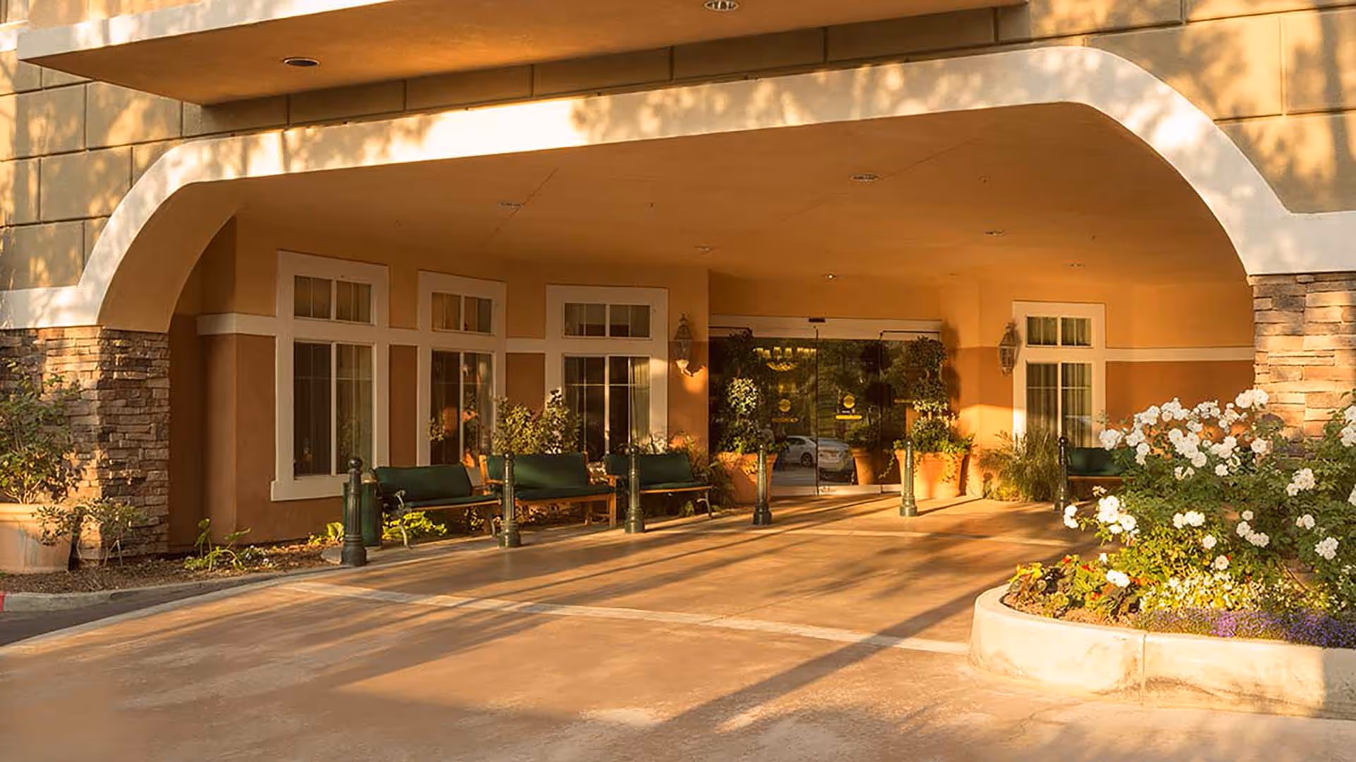 Covered entrance area of a building with stone and beige walls, several green benches, potted plants, and a flower bed with white flowers in the foreground. Glass doors lead inside the building.