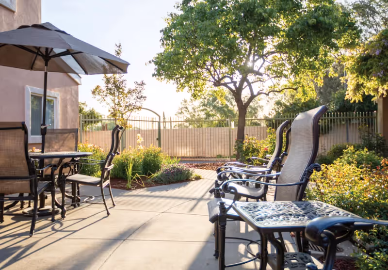 Sunlit outdoor patio with metal tables and chairs, an umbrella, and surrounding landscaping.
