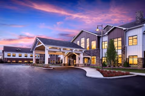 Exterior view of a senior living facility named Montage Mason at dusk, showing a large building with multiple windows illuminated from inside, a covered entrance with white pillars, a curved driveway, landscaped areas with small trees and shrubs, and a colorful sky with pink and purple clouds.