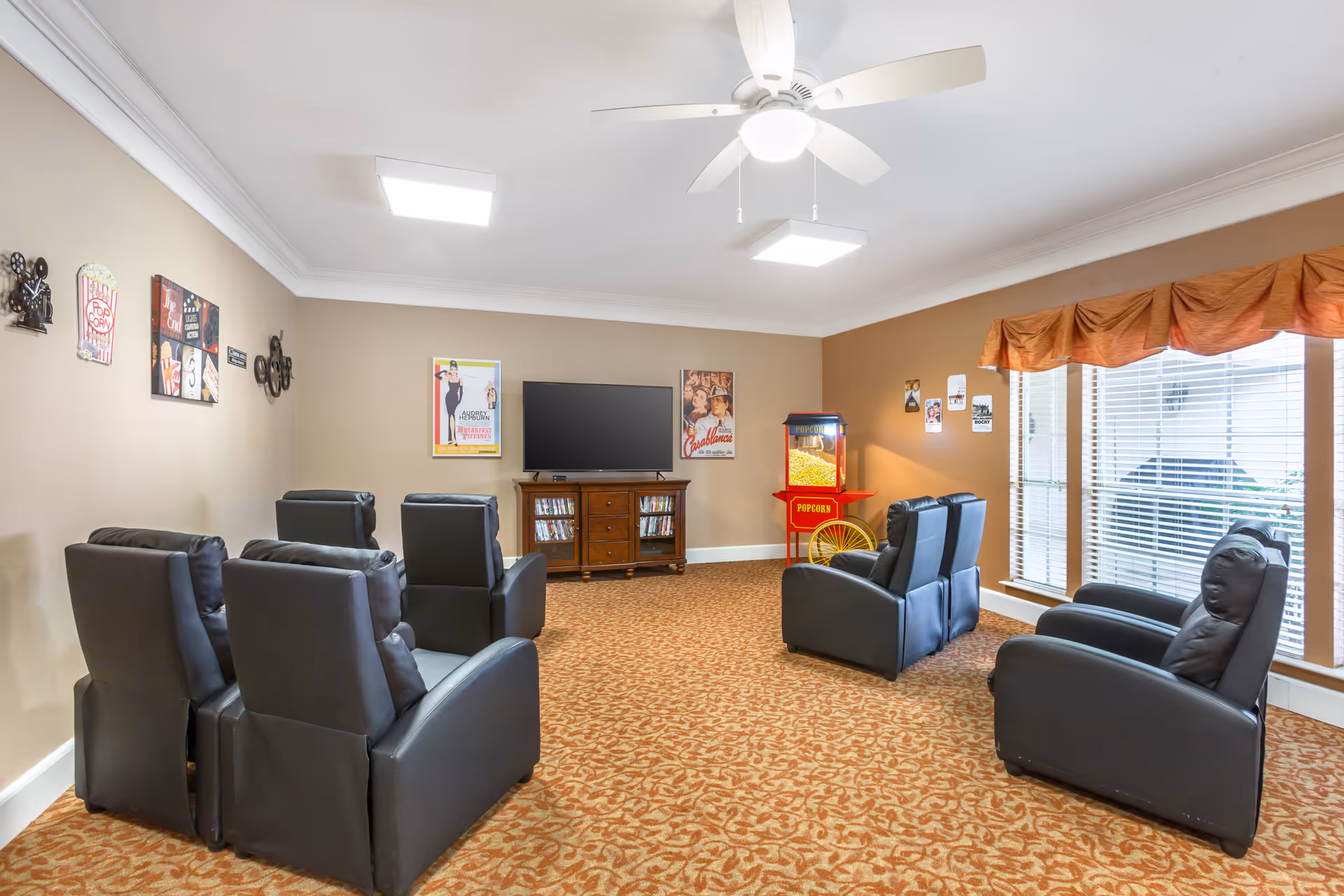A cozy entertainment room with six black recliner chairs arranged in two rows facing a flat-screen TV on a wooden stand. The room has beige walls decorated with vintage movie posters and a popcorn machine in the corner. Large windows with blinds and an orange valance allow natural light to enter. The ceiling has a white fan and recessed lighting, and the floor is covered with patterned carpet.